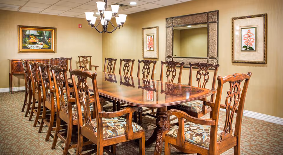 A formal dining room with a long polished wooden table surrounded by twelve ornate wooden chairs with floral upholstered seats. The room has beige walls adorned with framed artwork and a large decorative mirror. A chandelier with multiple lights hangs above the table.