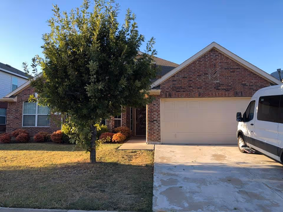 Front exterior view of a single-story brick house with a two-car garage, a tree in the front yard, and a white van parked in the driveway under a clear blue sky.