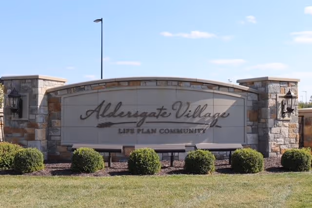 Stone and brick entrance sign for Aldersgate Village Life Plan Community with neatly trimmed bushes in front and a clear blue sky in the background.