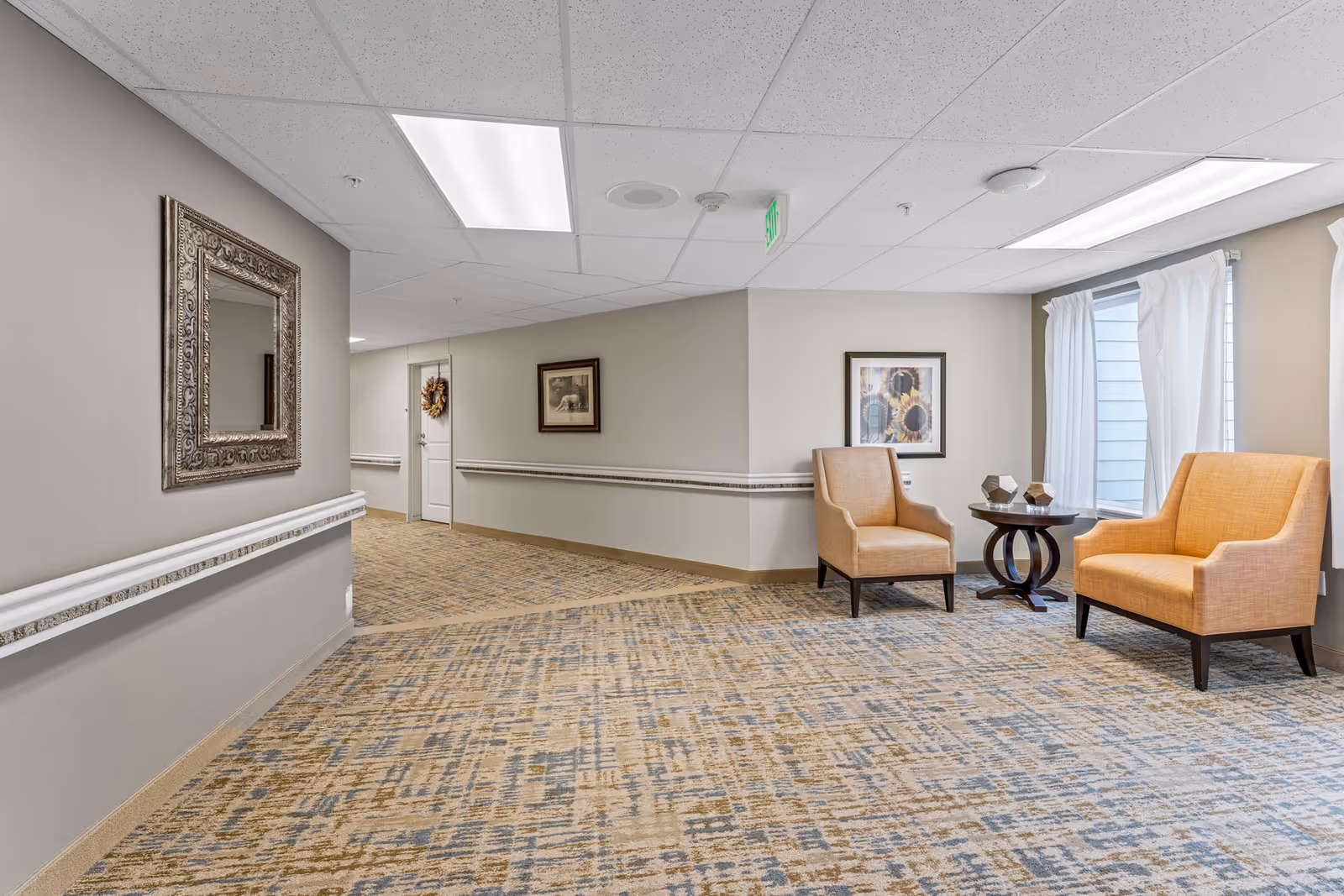 A well-lit hallway in a senior living facility featuring beige walls and patterned carpet. There are two orange upholstered armchairs with a small round wooden table between them, positioned near a window with white curtains. The walls are decorated with framed artwork and a large ornate mirror. The hallway has handrails along the walls and a door at the end with a wreath hanging on it.
