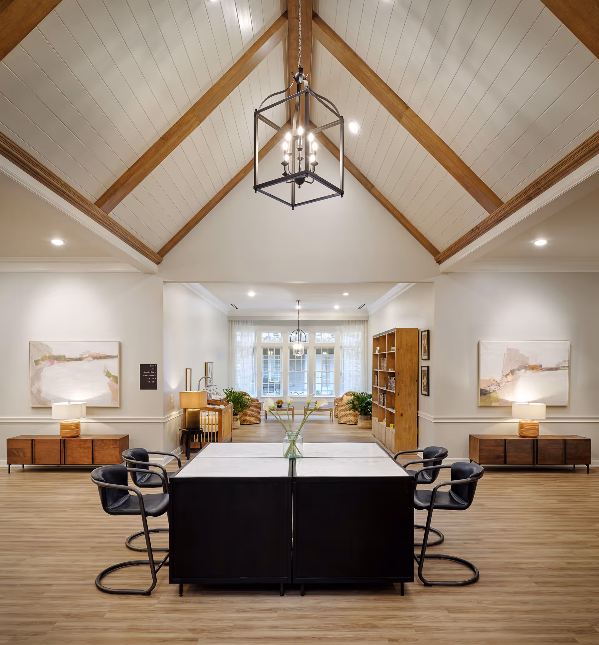 Spacious senior living common area with a vaulted wood-beamed ceiling, a central table with chairs, pendant lighting, and a seating area in the background.