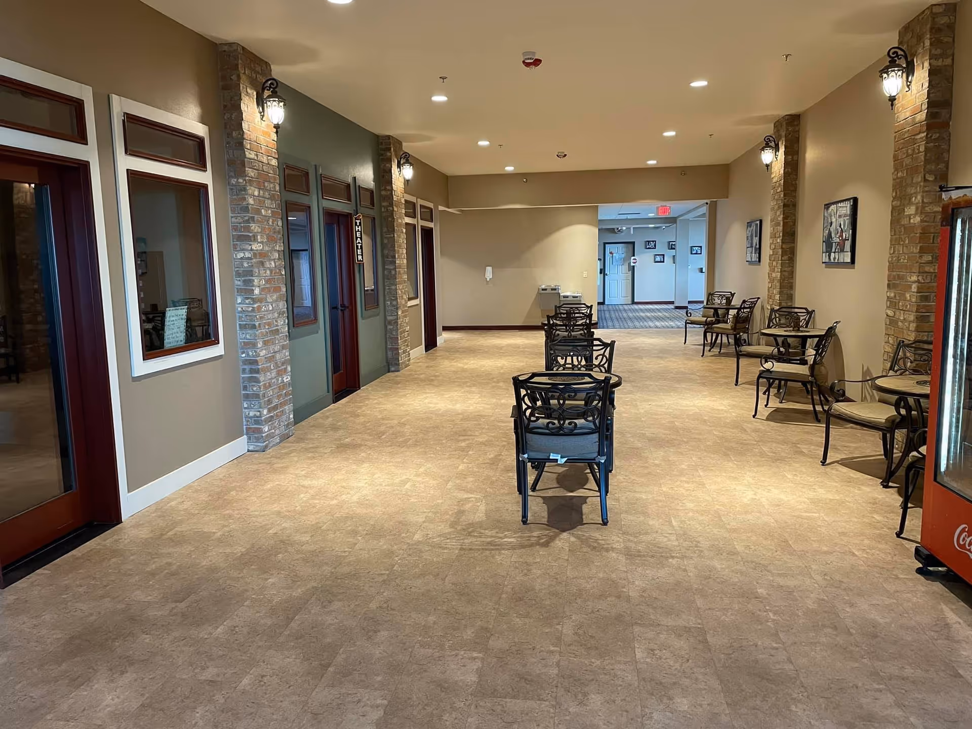 Wide indoor common corridor in an assisted living facility with rows of metal chairs and small tables, brick columns, and wall sconces.