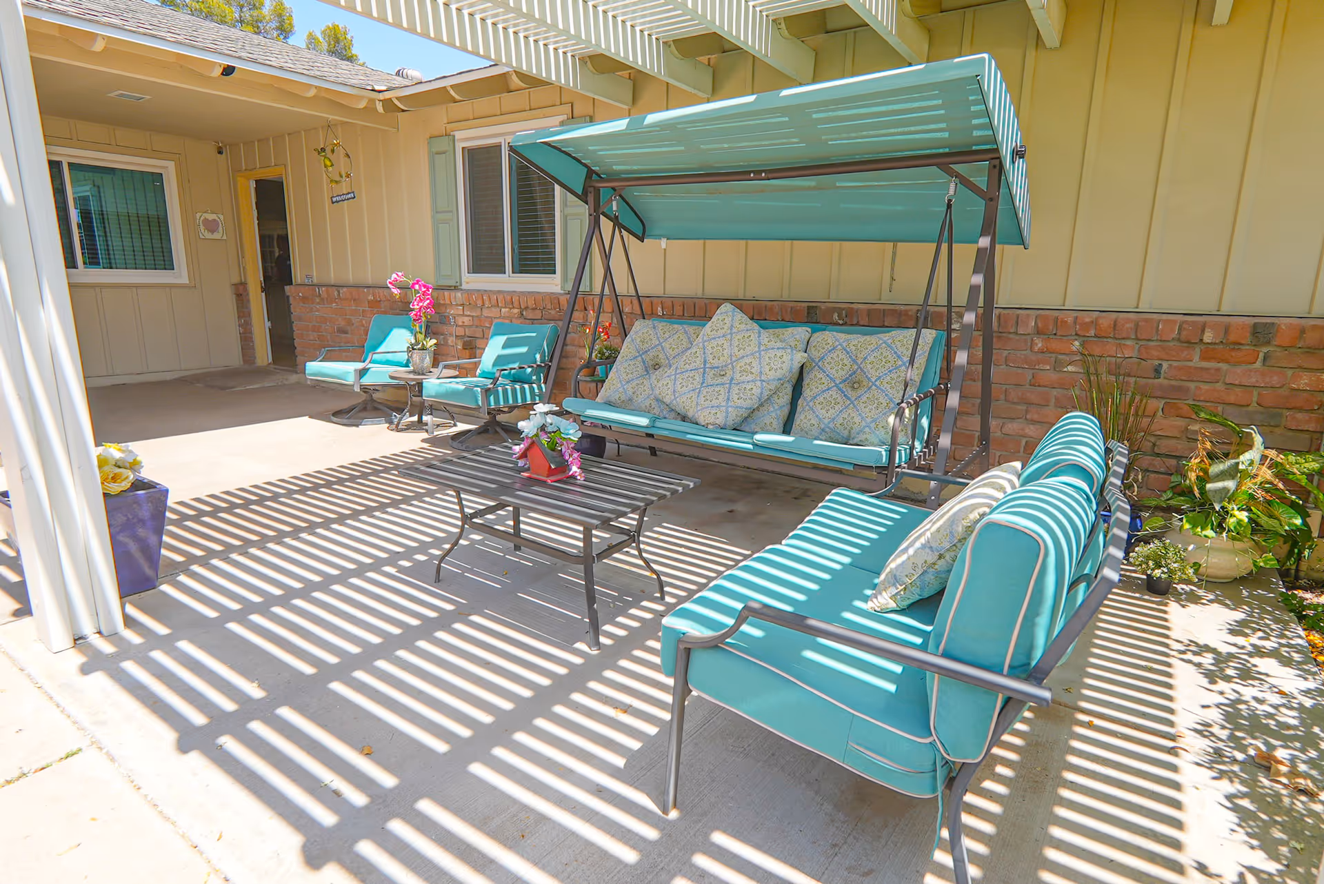 Outdoor patio area with teal cushioned seating including a swing, chairs, and a loveseat under a pergola casting striped shadows on the concrete floor. There are potted plants and a small table with flowers in the center.