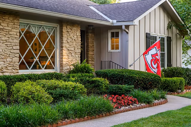 Exterior view of a single-story home with stone and siding walls, a large window with diamond-shaped panes, a Kansas City Chiefs flag mounted near the entrance, and well-maintained landscaping including bushes, grass, and flowers along a paved walkway.