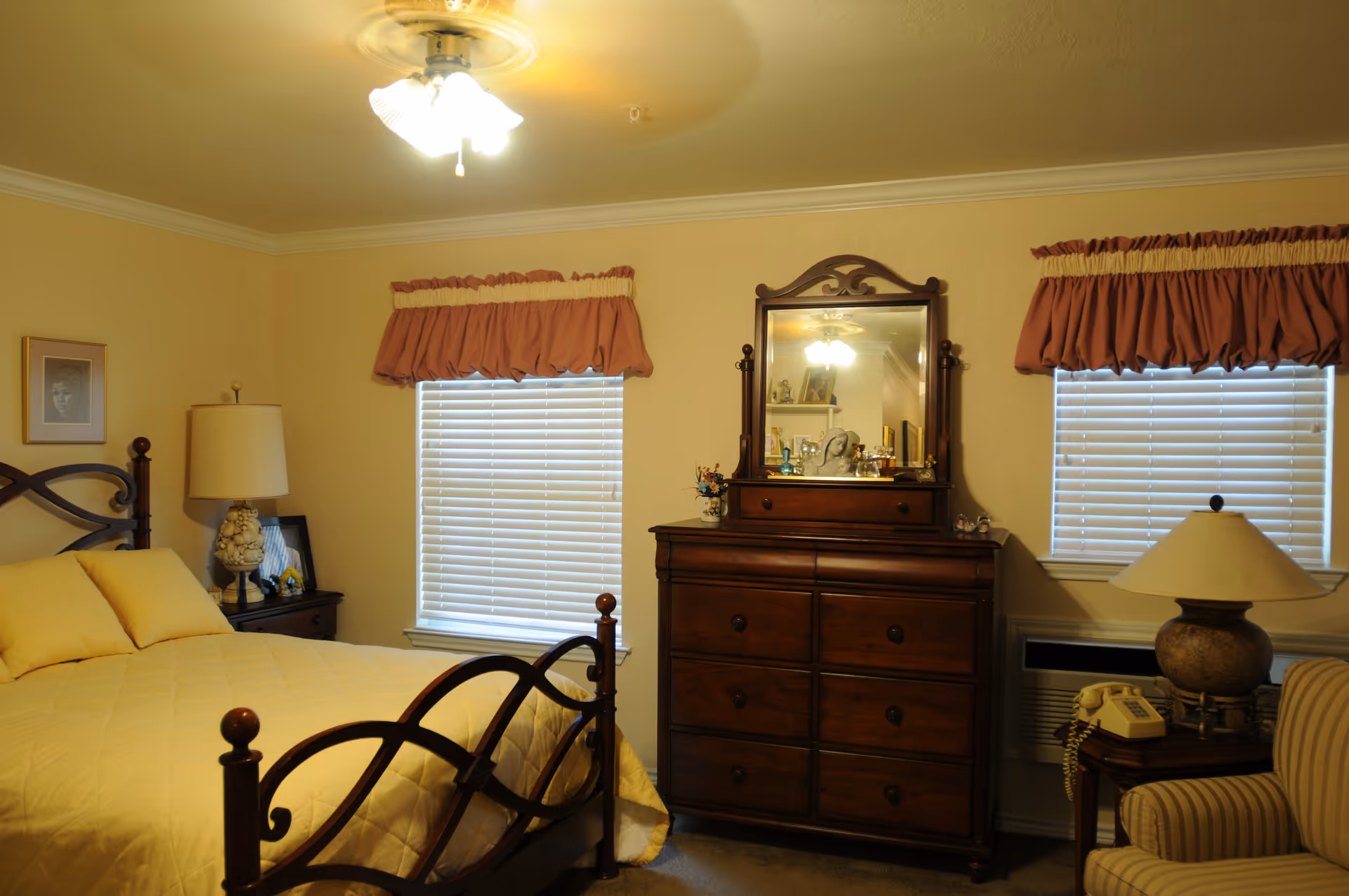 A cozy bedroom with a wooden bed frame and yellow bedding, two windows with blinds and red valances, a wooden dresser with a mirror, a nightstand with a lamp and framed photo, and an armchair with a side table holding a lamp and a vintage telephone.