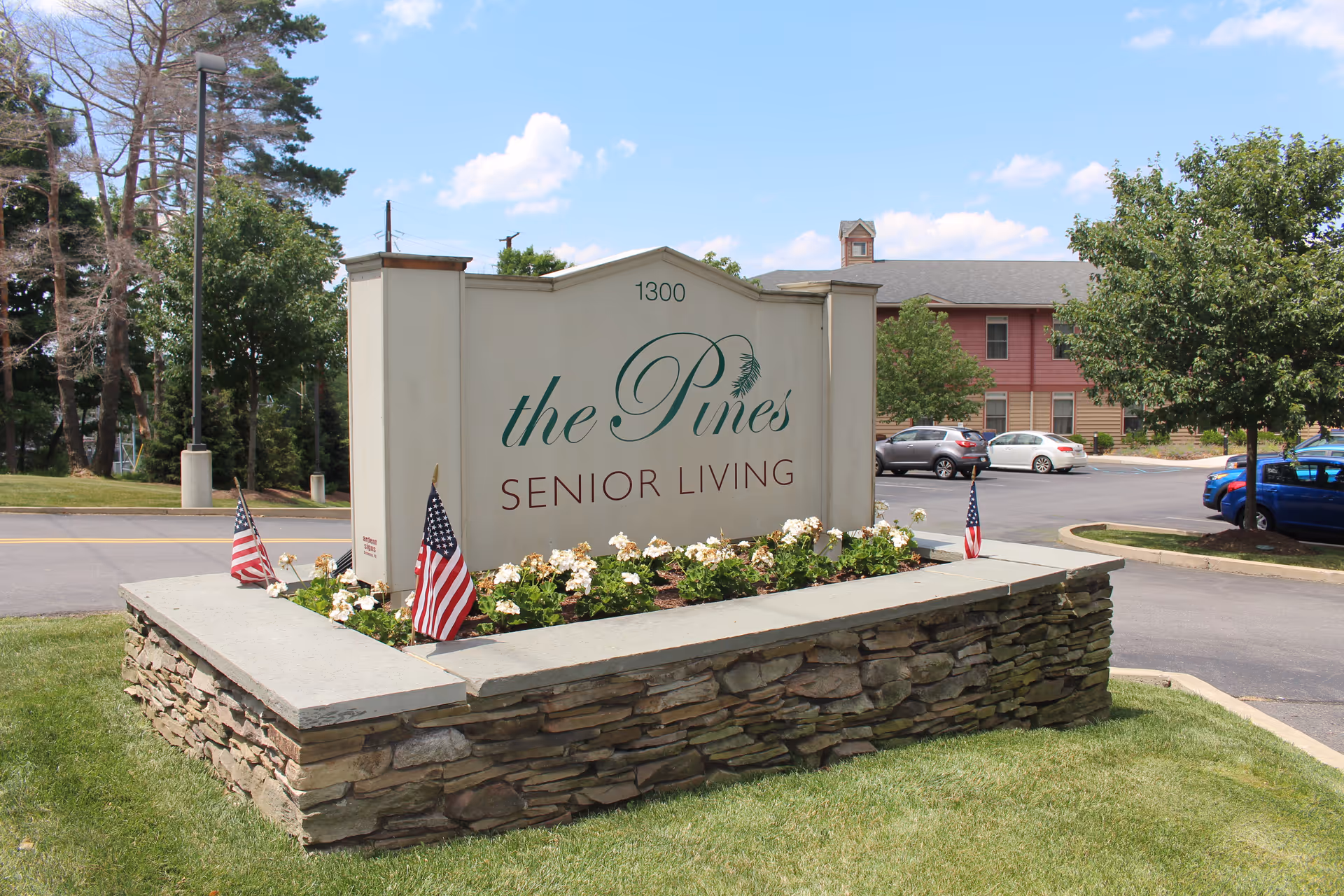Entrance sign in a stone planter reading 'the Pines SENIOR LIVING' with the facility building and parked cars behind it.