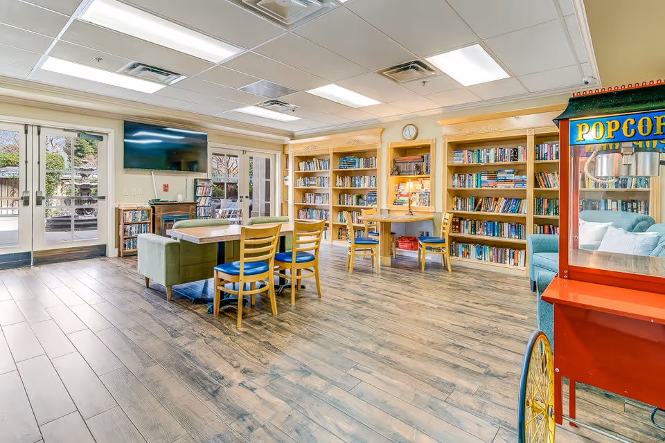 Bright community library/lounge with bookshelves, tables and chairs, a wall-mounted TV, and a popcorn cart.