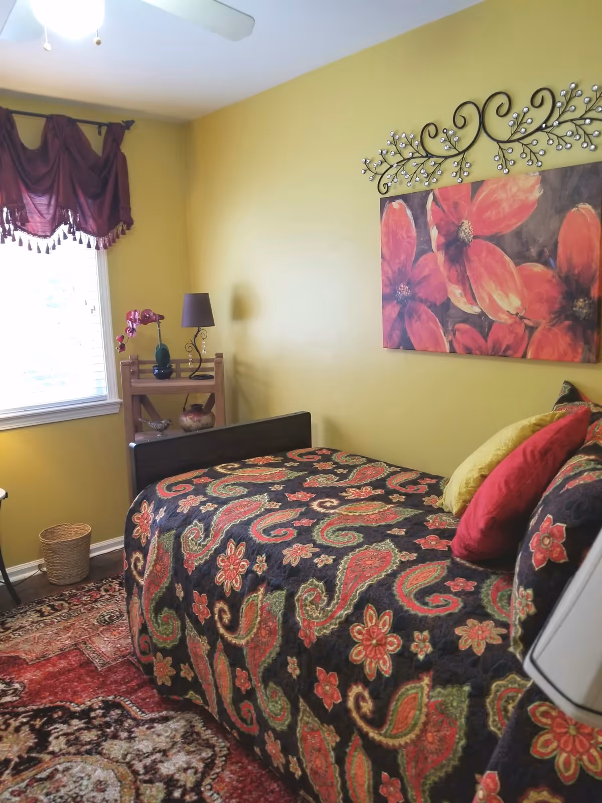 A cozy bedroom with yellow walls featuring a bed covered with a colorful floral and paisley patterned quilt. The bed has multiple pillows in red, yellow, and black. Above the bed is a large painting of red flowers with a decorative metal wall art piece above it. To the left, there is a window with a burgundy valance, a small wooden shelf with a lamp, a vase with flowers, and decorative items. A patterned rug covers the floor.