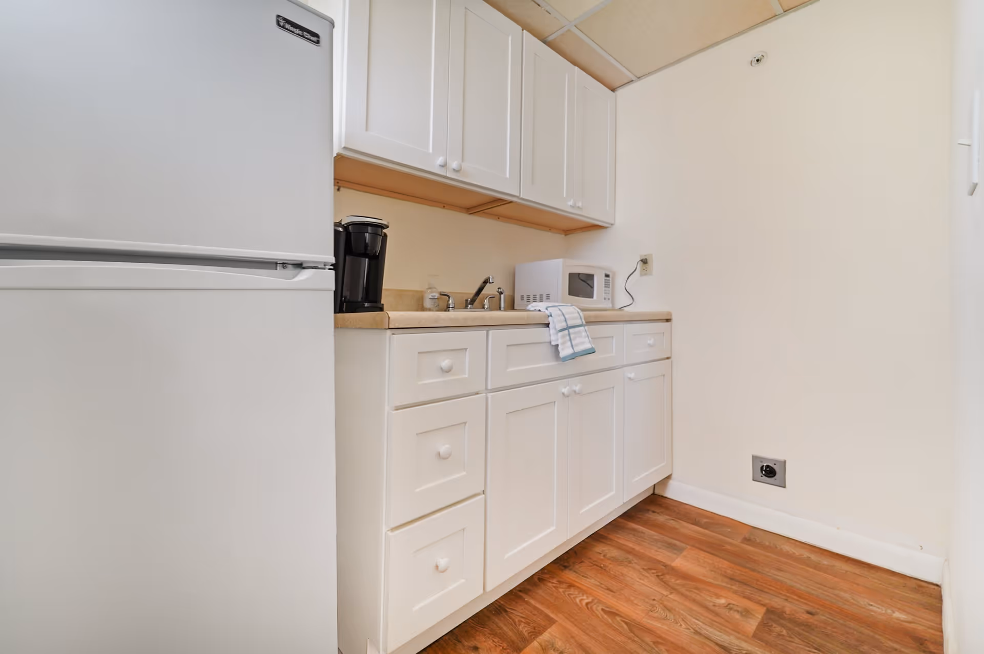 A small kitchen area with white cabinets, a beige countertop, a white refrigerator, a black coffee maker, a microwave, and a sink with a faucet. The floor is wooden, and the walls are painted white.