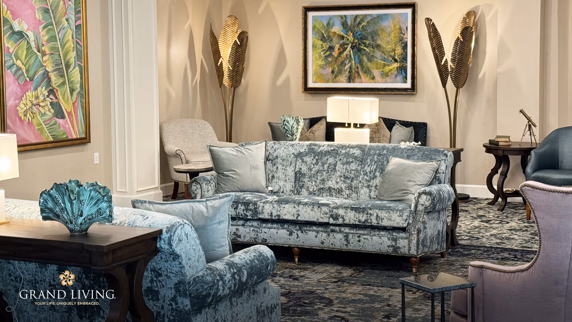 A cozy living room area with plush blue velvet sofas, a patterned carpet, decorative pillows, a wooden side table with a blue decorative piece, a lamp, and framed artwork on the walls. The room features warm lighting and elegant decor including gold leaf-shaped floor lamps.