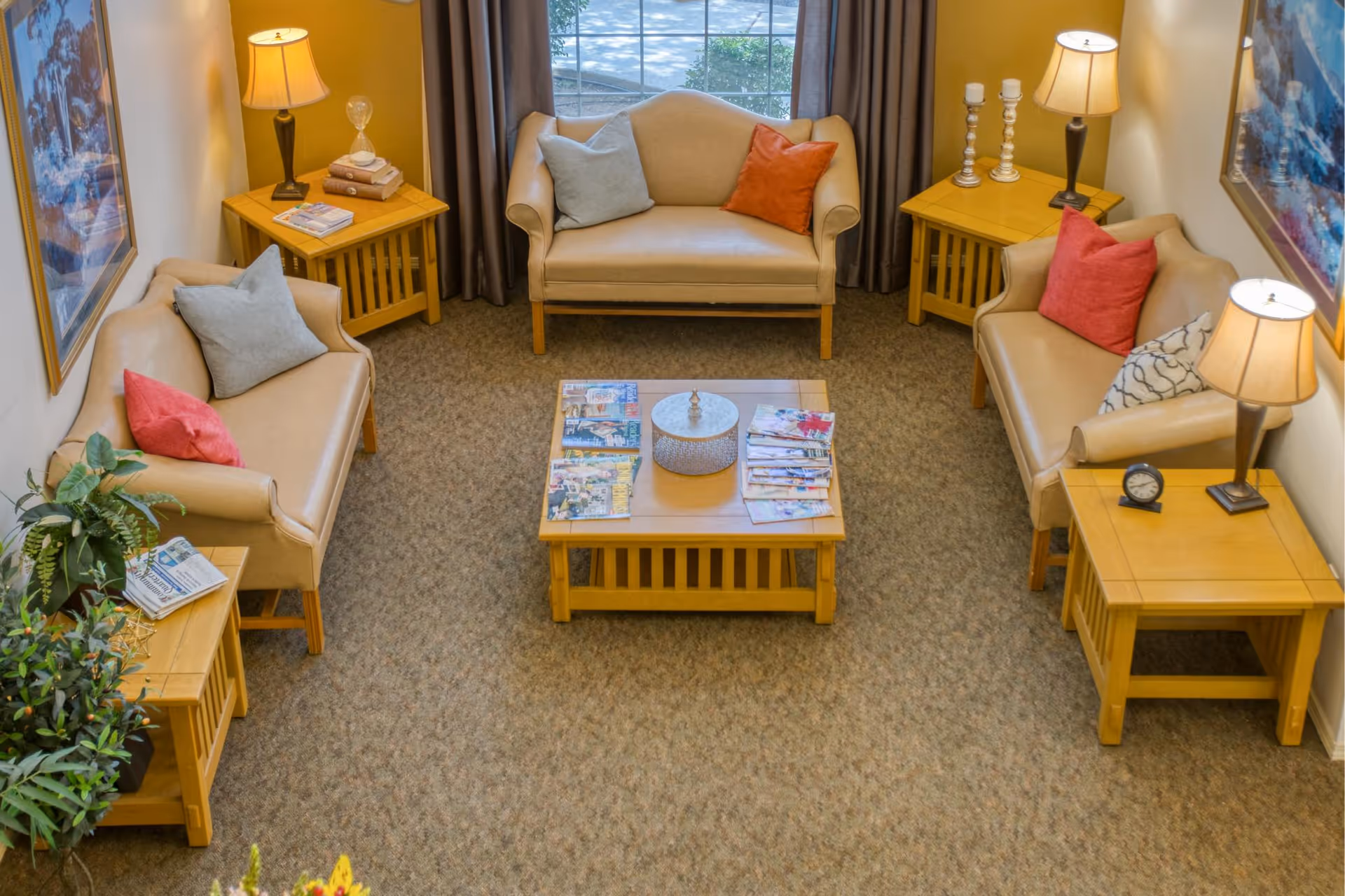 A cozy senior living lounge with three beige sofas, wooden side tables and a central coffee table topped with magazines.
