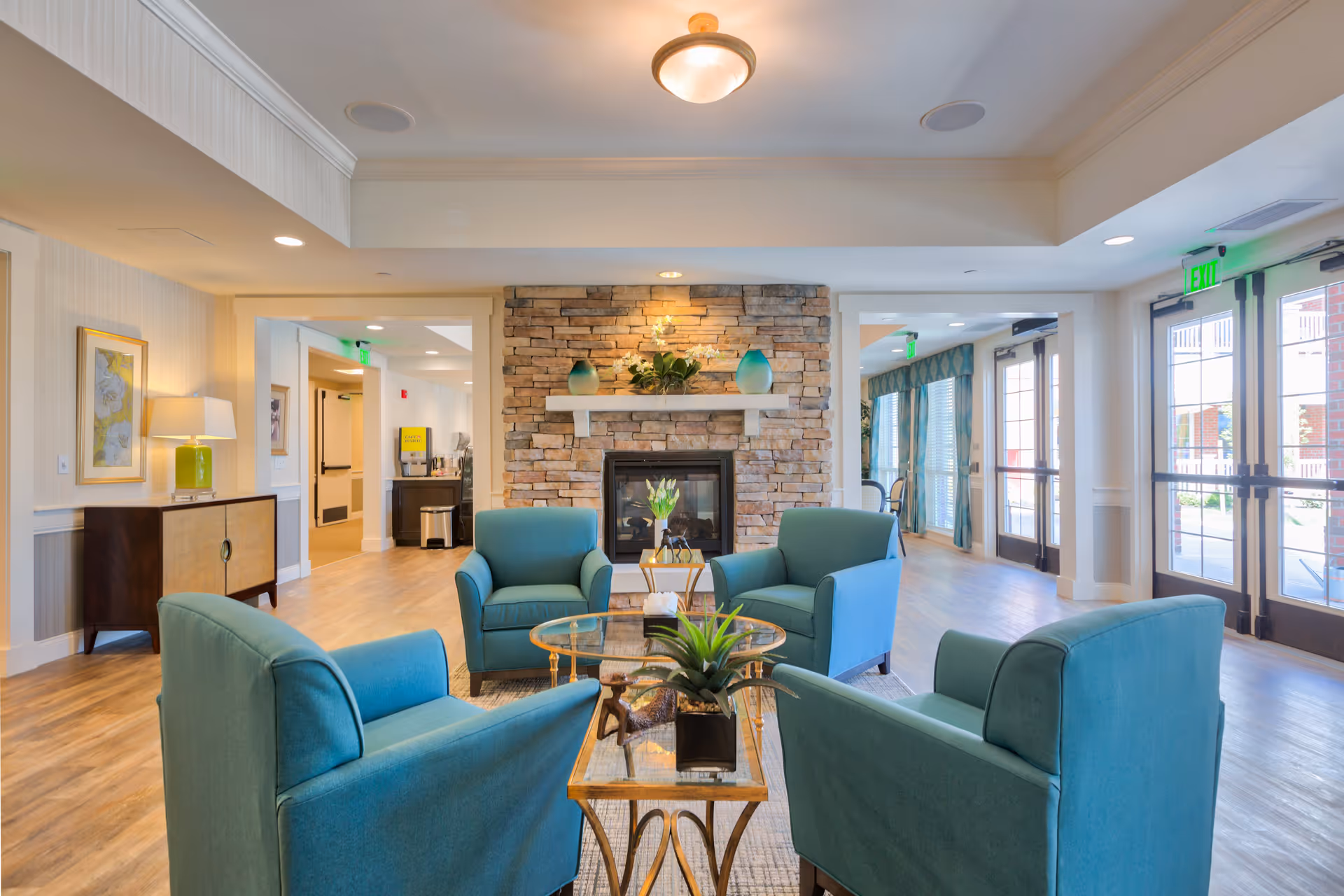 A bright and inviting common area with four teal armchairs arranged around a glass coffee table. The room features a stone fireplace with decorative vases and a plant on the mantel. Large windows and glass doors allow natural light to fill the space, and light wood flooring complements the neutral walls and ceiling.