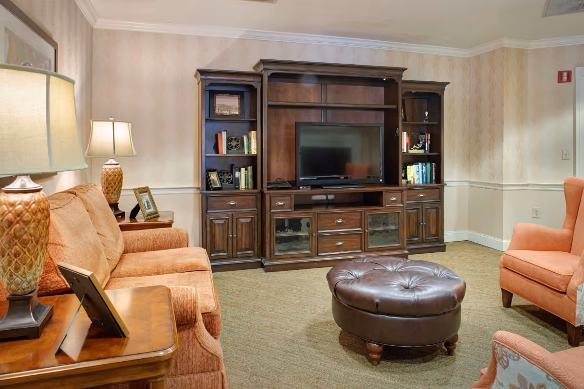 Comfortable living room with upholstered sofas and chairs, a round leather ottoman, lamps, and a large wooden entertainment center with a TV.