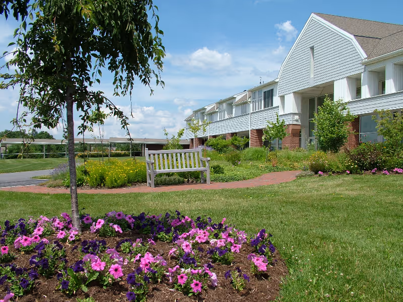 Outdoor garden area with a wooden bench on a brick pathway, surrounded by green grass, colorful flowers, and small trees. A large building with white siding and brick accents is visible in the background under a partly cloudy blue sky.