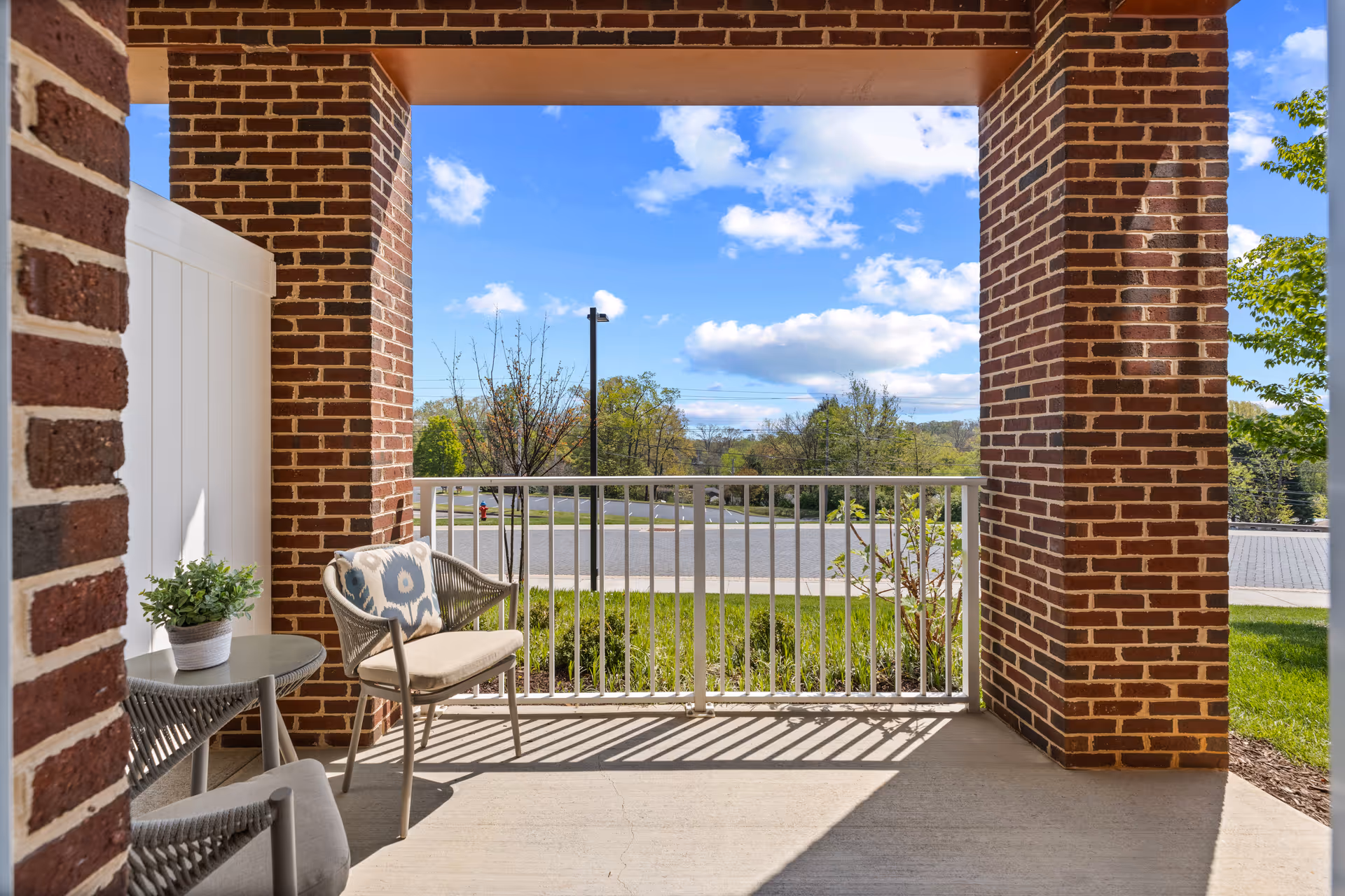 A covered outdoor patio area with brick pillars and a white railing overlooking a street and greenery. The patio has two chairs with cushions and a small round table with a potted plant on it. The sky is partly cloudy and blue.