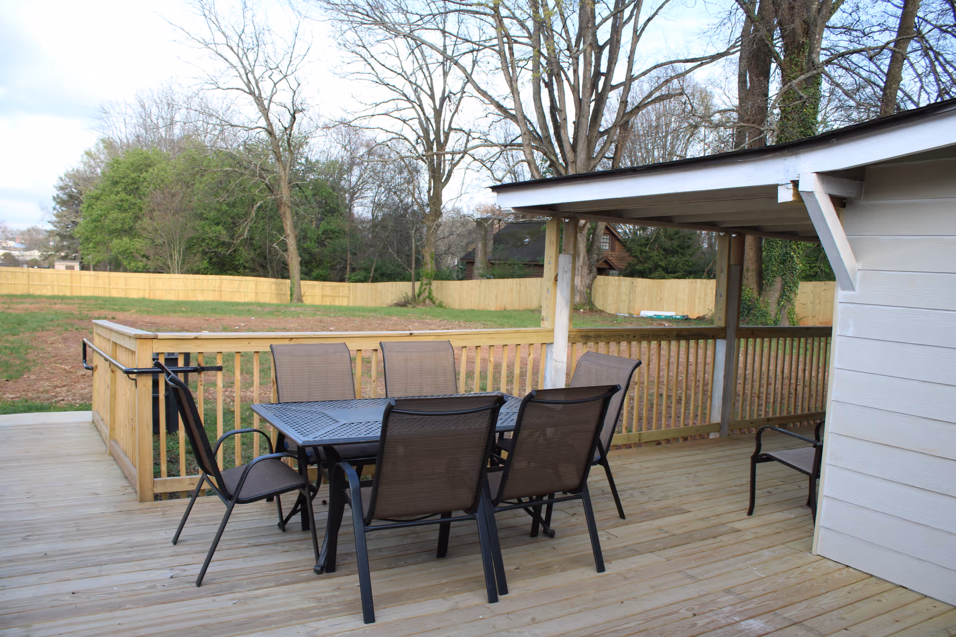 Outdoor wooden deck area with a black metal table and six matching chairs. The deck has a wooden railing and is partially covered by a roof. In the background, there is a grassy yard with trees and a wooden fence.