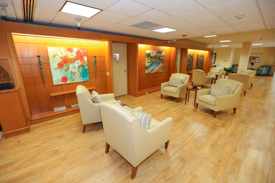 A spacious and well-lit lounge area with several beige armchairs arranged around small wooden tables on a light wood floor. The walls feature wooden paneling with colorful floral and nature-themed paintings, and the ceiling has recessed lighting. The overall atmosphere is warm and inviting.