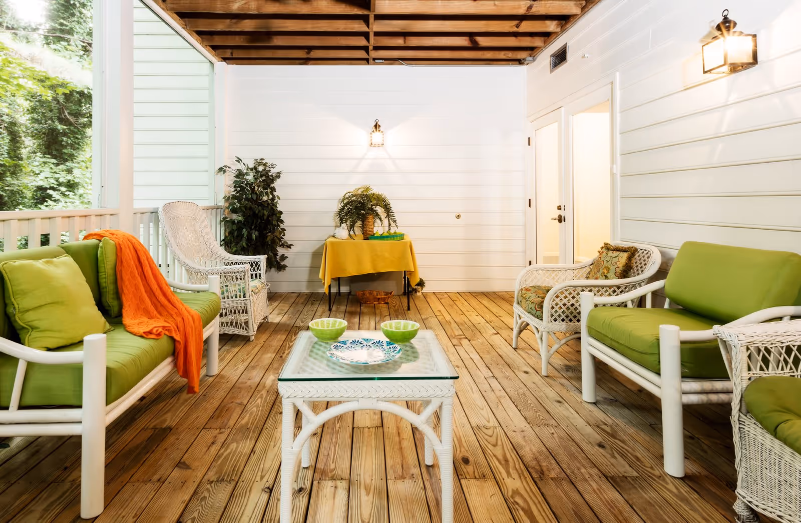 Covered outdoor patio area with wooden floor and ceiling beams, furnished with white wicker chairs and sofas with green cushions, a glass-top wicker coffee table with decorative bowls, a small table with a yellow tablecloth and plants, and a wall-mounted lantern light.