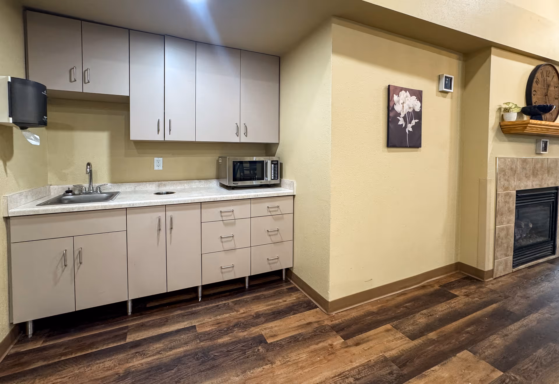 Interior view of a kitchenette area with beige cabinets, a countertop with a sink, a microwave, and a paper towel dispenser mounted on the wall. The room has wood flooring, beige walls, and a partial view of a fireplace with a clock and decorative items on the mantel.