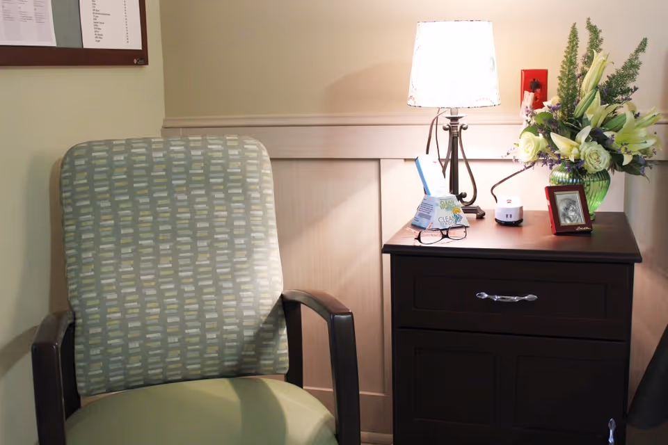 A cozy corner in a room featuring a patterned armchair next to a dark wooden nightstand. The nightstand holds a lit table lamp, a green vase with white and purple flowers, a small framed photo, a pair of glasses, and a few small items. The wall behind is light-colored with a bulletin board partially visible on the left.
