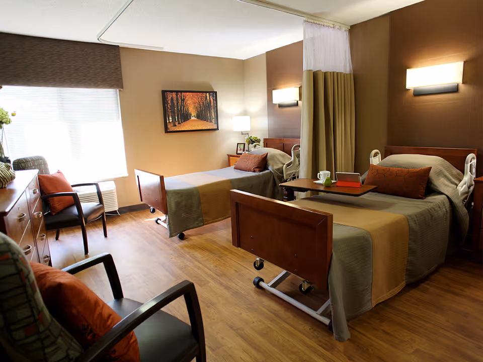 A well-lit room with two hospital-style beds, each with brown and beige bedding and orange pillows. There is a wooden floor, two chairs with orange cushions, a wooden dresser, and a window with blinds. A painting of a tree-lined path with autumn leaves hangs on the wall. A privacy curtain is partially drawn between the beds, and there are wall-mounted lights above each bed.