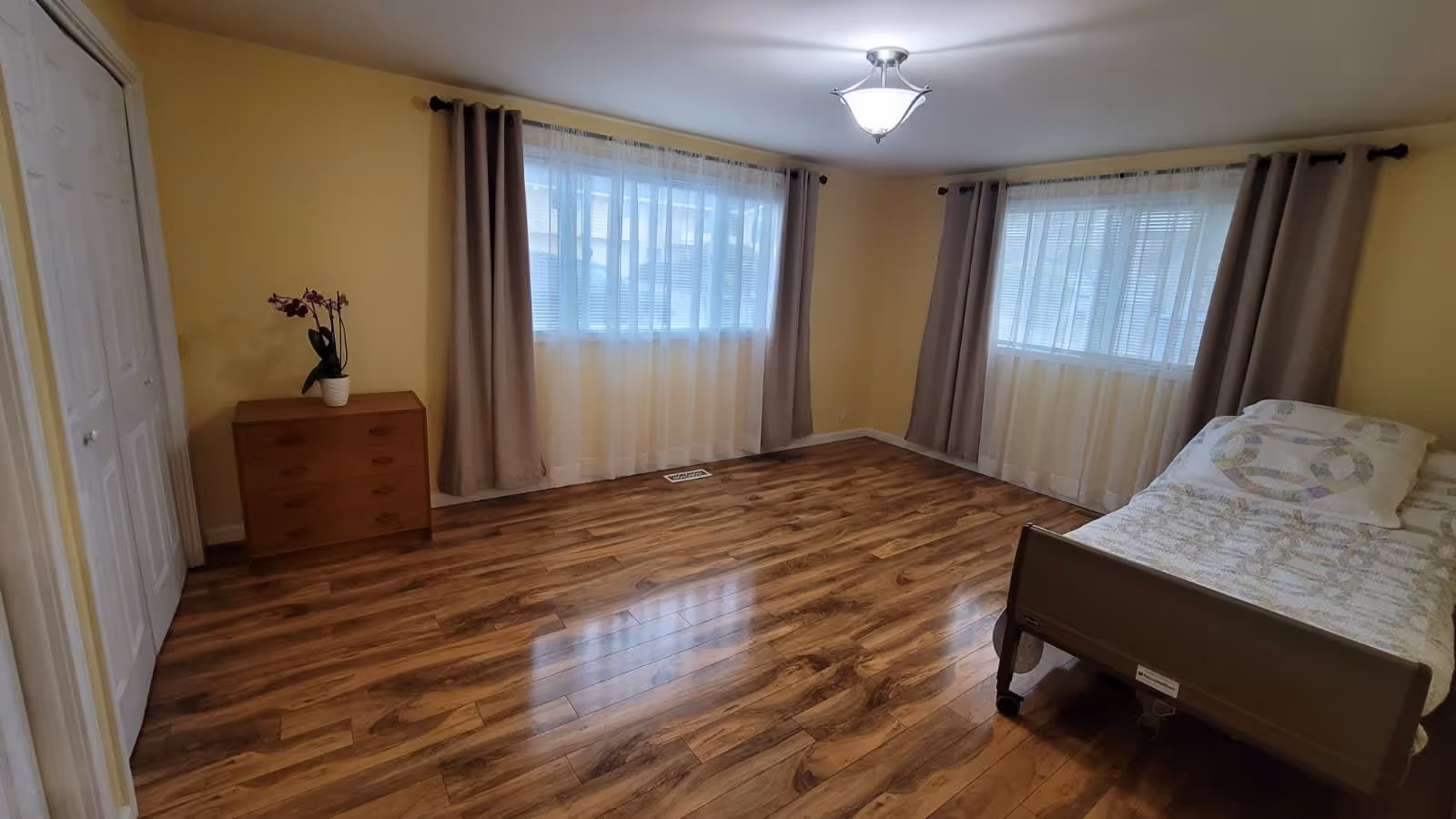 A spacious bedroom with wooden flooring, two large windows with sheer white curtains and beige drapes, a single bed with a patterned quilt and pillow, a wooden dresser with a potted orchid, and a ceiling light fixture.