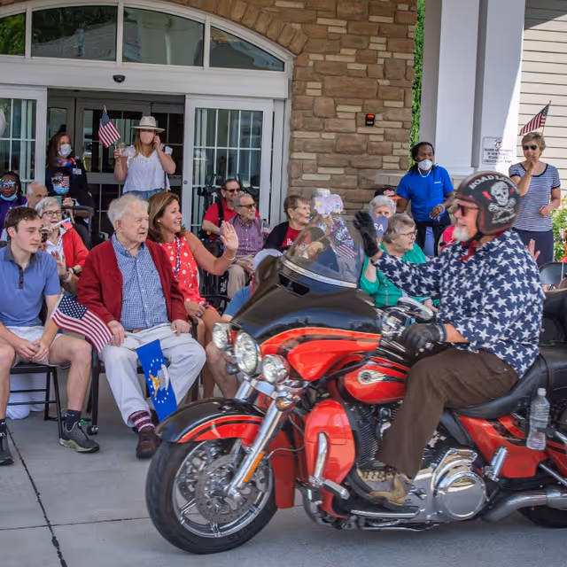 A group of elderly people and some younger individuals sitting outside a building, watching and waving at a man riding a red and black motorcycle. The man on the motorcycle is wearing a star-patterned shirt, a helmet with skull designs, and gloves. Some people in the crowd are holding small American flags, and a few are wearing masks. The building behind them has stone and white pillars.