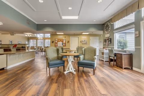 A spacious, well-lit common area with wooden flooring featuring two green upholstered armchairs and a small round table in the center. To the right, there is a window with blinds, a wall clock, and a desk with a chair. In the background, there is a dining area with tables and chairs, and a counter with a mirror above it on the left side.