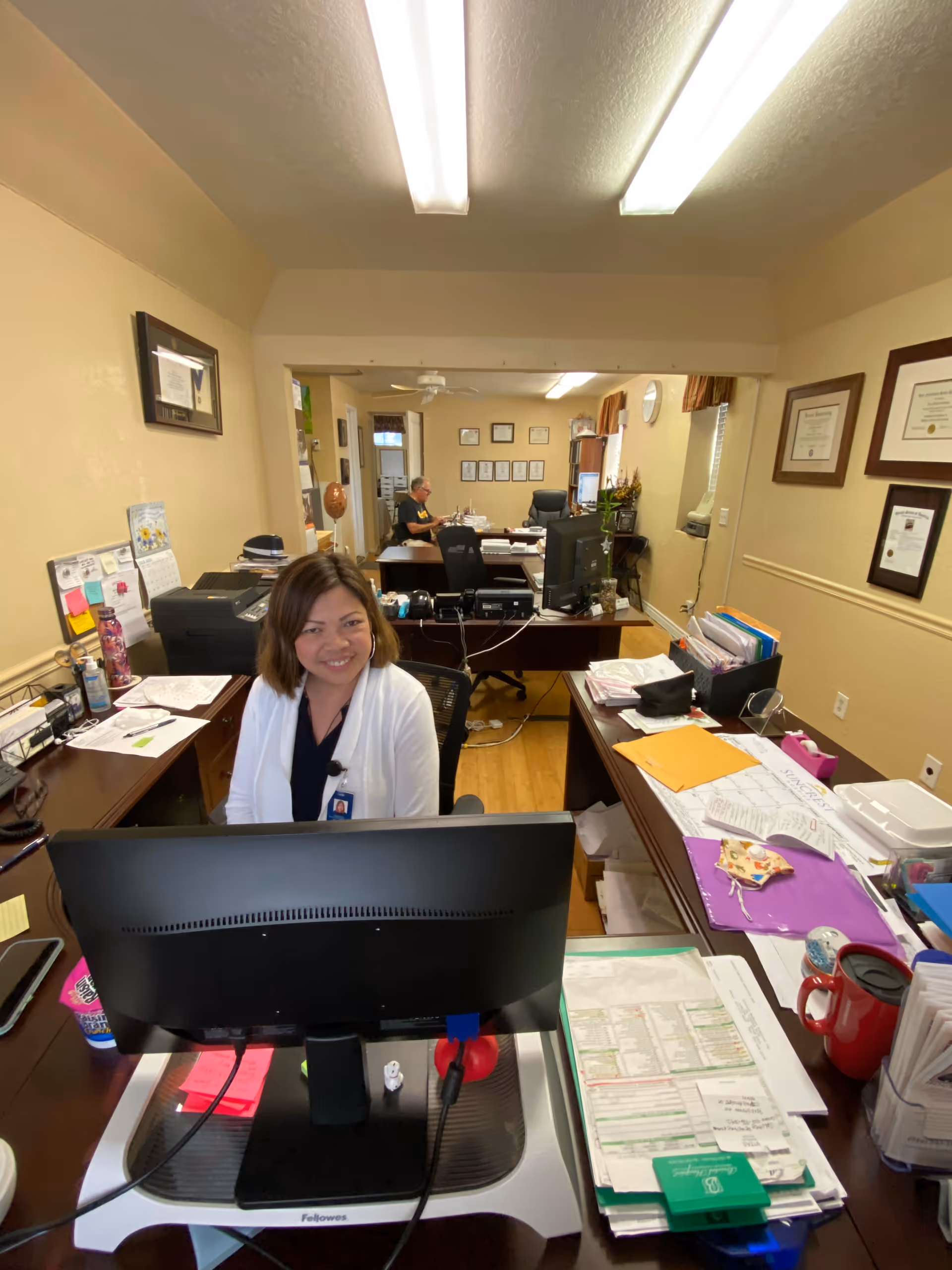 A smiling staff member sits at a desk behind a computer in a busy administrative office with papers, monitors, and framed certificates on the walls.