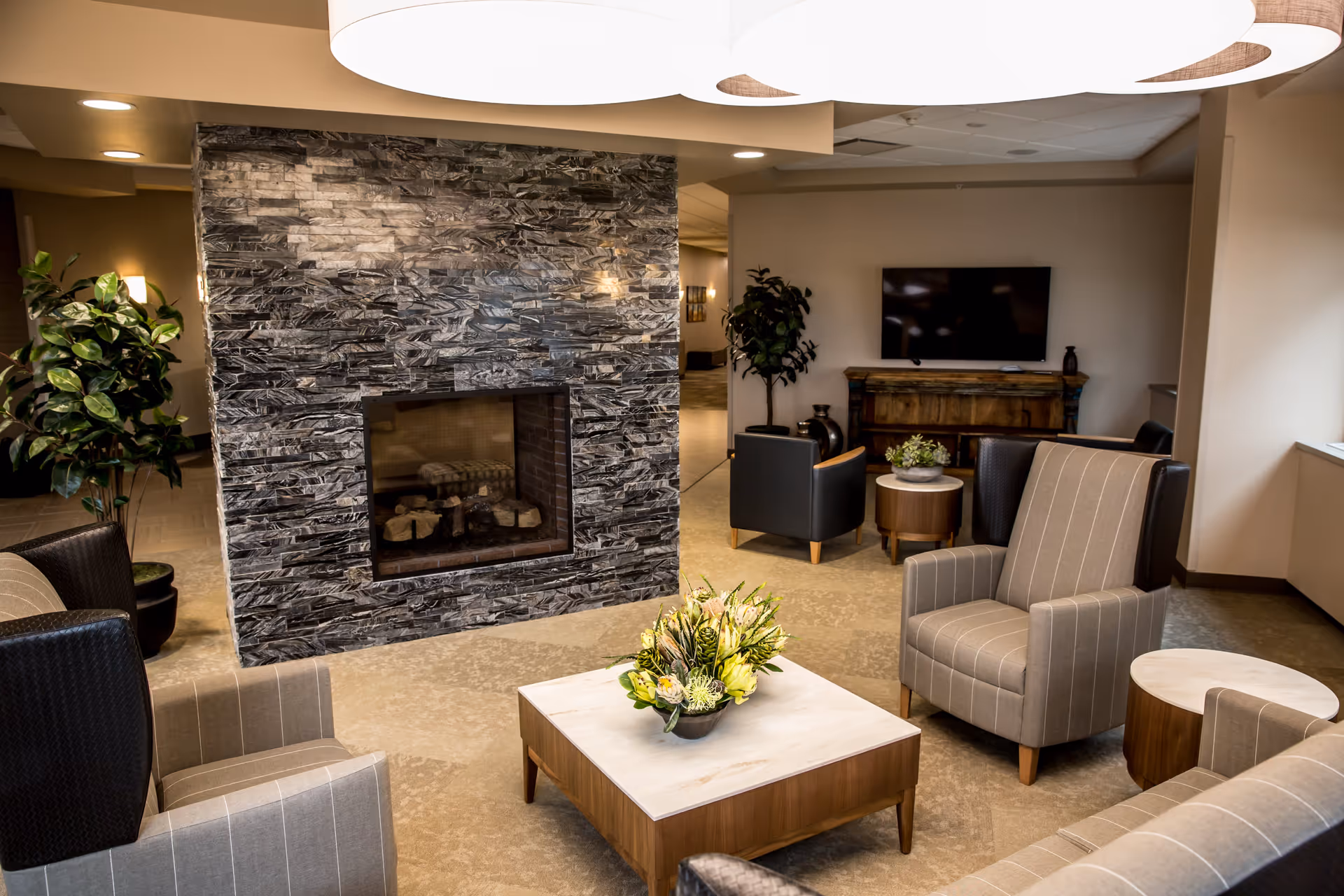 A cozy living room area in a senior living facility featuring a modern stone fireplace, several upholstered armchairs with striped fabric, a wooden coffee table with a floral arrangement, a flat-screen TV mounted on the wall, and decorative plants.