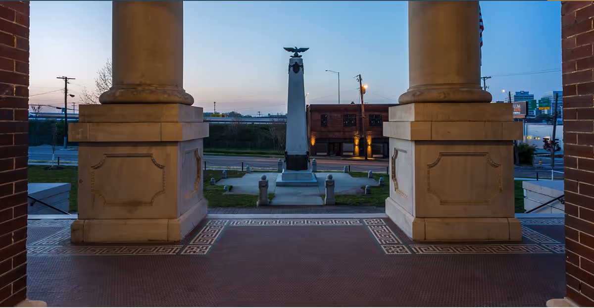 View from a building's tiled portico framed by two large stone columns overlooking an obelisk monument and street at dusk.