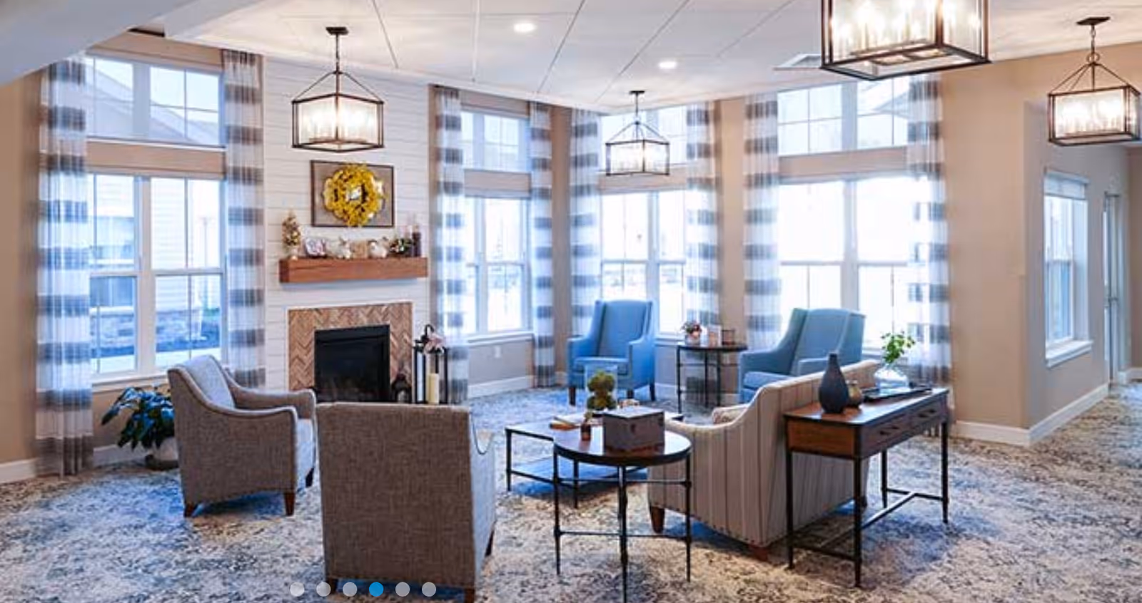 A bright and spacious living room with large windows covered by striped curtains, a fireplace with a wooden mantel decorated with a yellow wreath and small ornaments, two blue armchairs, two gray armchairs, a striped sofa, and several tables with decorative items. The room has a patterned carpet and modern ceiling lights.