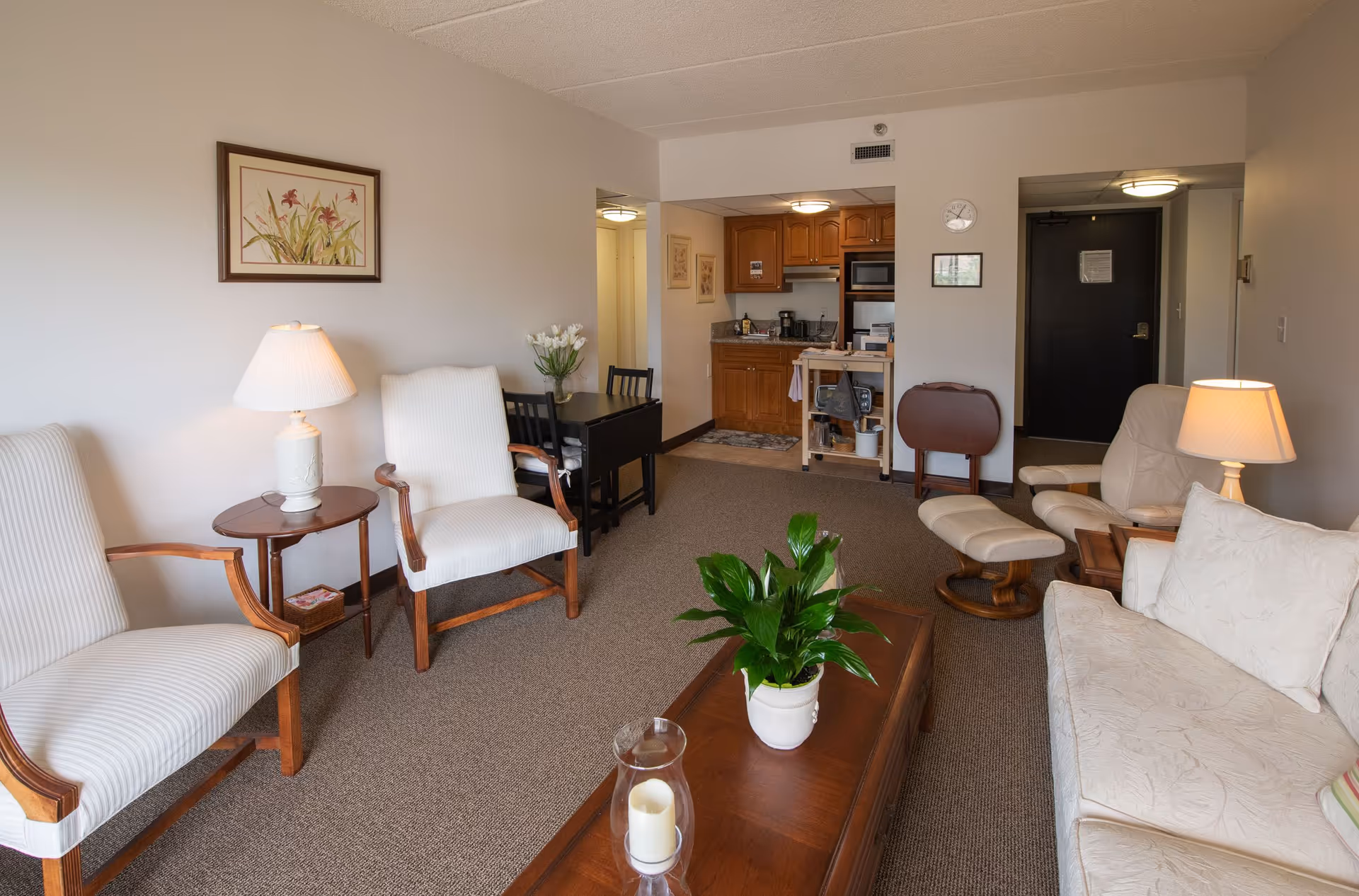 A cozy living room area in a senior living facility with a beige sofa, two white armchairs, a wooden coffee table with a potted plant and a candle, a small dining table with chairs, and a kitchenette in the background. The room is carpeted and decorated with a framed floral painting and table lamps providing warm lighting.