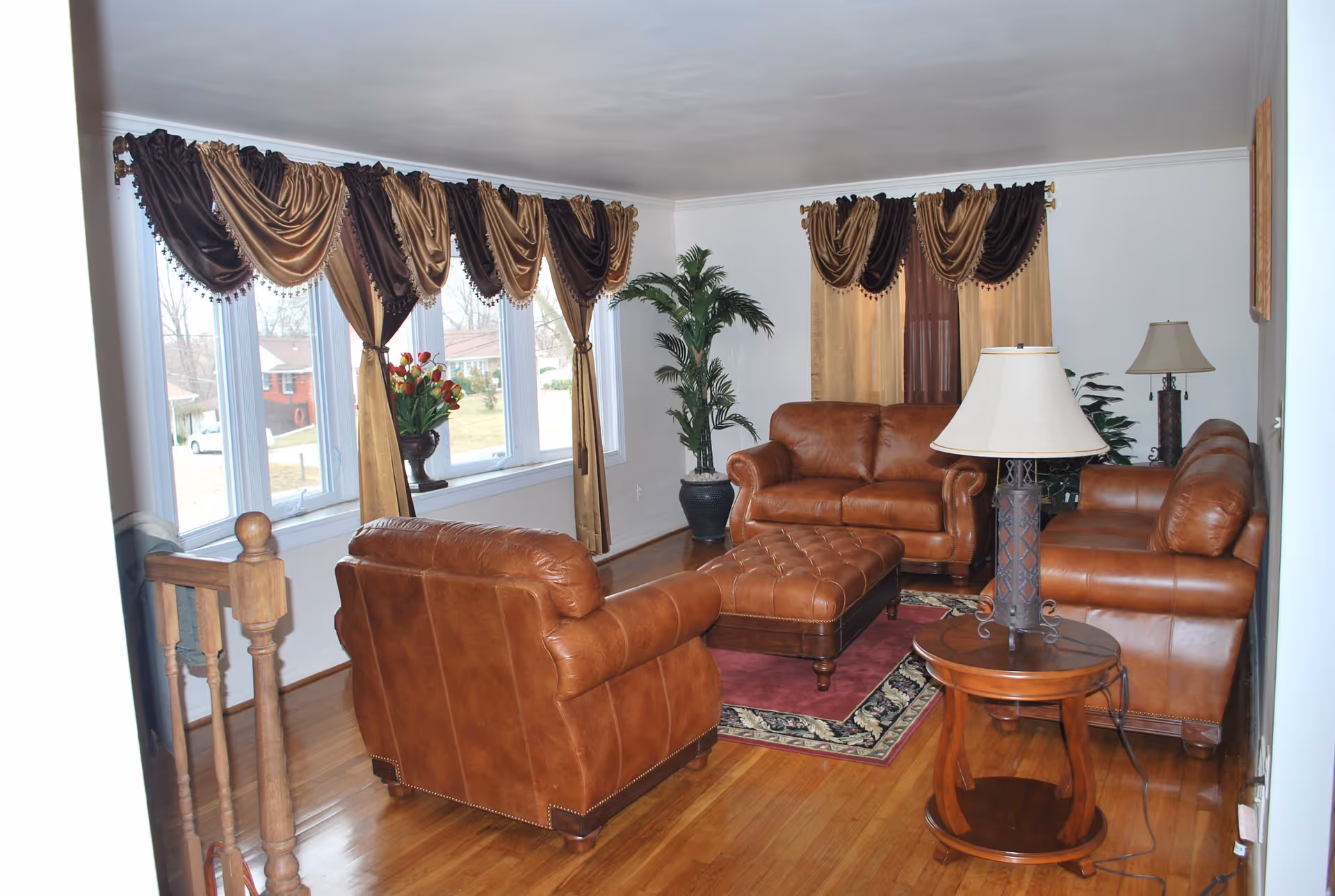 A living room with three brown leather sofas arranged around a matching brown leather ottoman on a decorative rug. There are two table lamps on wooden side tables, a large potted plant in the corner, and windows with brown and gold drapes letting in natural light.