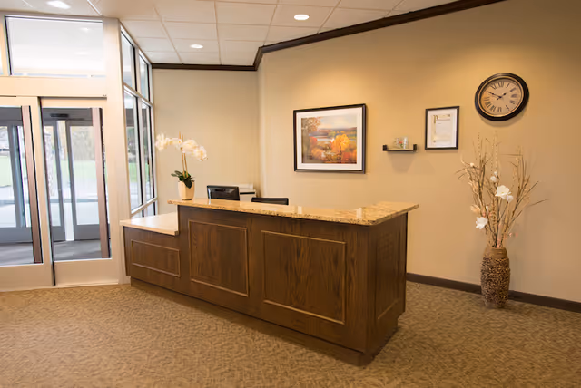Reception area with a wooden front desk topped with granite, a vase with white flowers on the left side, a wall clock, framed artwork, and a decorative vase with dried branches on the right side. Glass doors and windows are visible in the background.