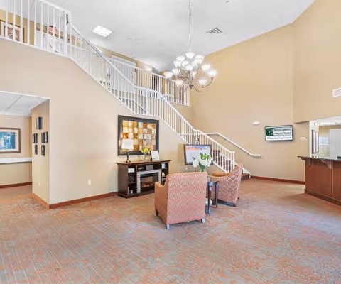 A spacious interior common area with beige walls and a high ceiling. There is a staircase with white railings leading to an upper floor. Two patterned armchairs and a small round table with flowers are placed on a carpeted floor. A wooden console with a lamp and decorative items is against the wall. A chandelier hangs from the ceiling, and a reception desk is visible on the right side. A sign on the wall reads 'Bentley Commons at Paragon Village'.