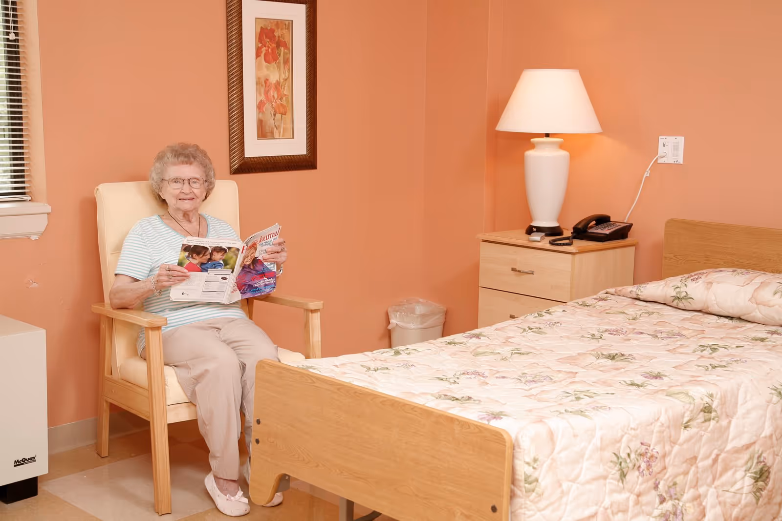 An elderly woman with glasses sitting in a wooden armchair with cream cushions, reading a magazine in a room with peach-colored walls. Next to her is a bed with a floral quilt, a wooden nightstand with a white lamp and a telephone, and a framed floral painting on the wall.
