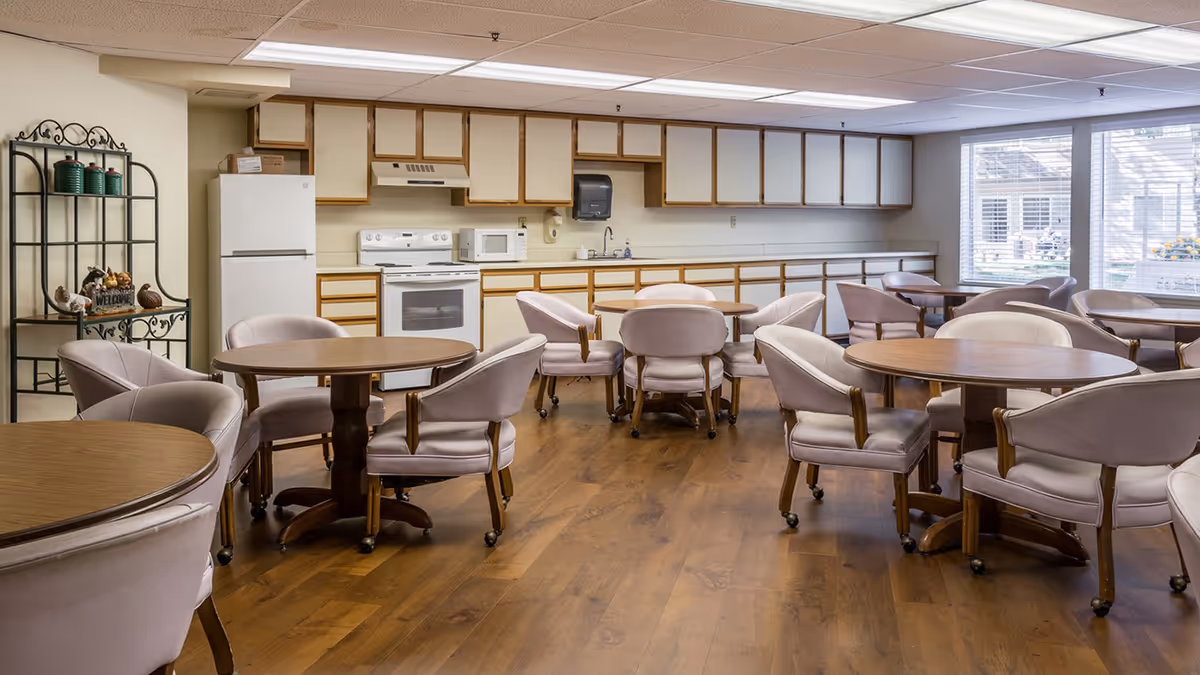 A communal dining area with several round wooden tables surrounded by cushioned chairs on wheels. The room features a kitchen area along the back wall with white cabinets, a refrigerator, stove, microwave, and a paper towel dispenser. Large windows on the right side let in natural light, and a decorative metal shelf with green canisters and a welcome sign is on the left.