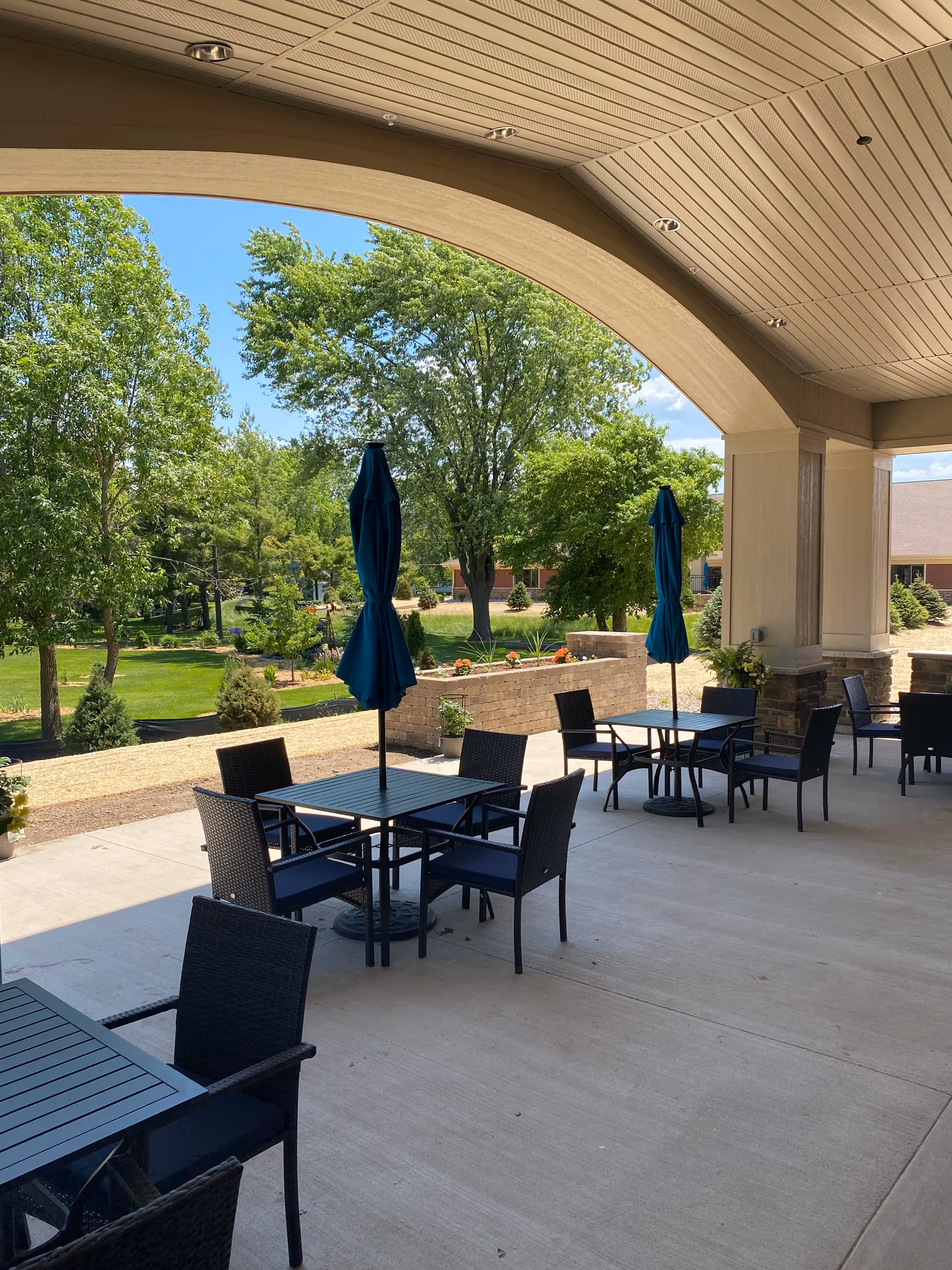 Covered outdoor patio area with several black tables and chairs, each table having a closed blue umbrella. The patio overlooks a green lawn with trees and shrubs under a clear blue sky.