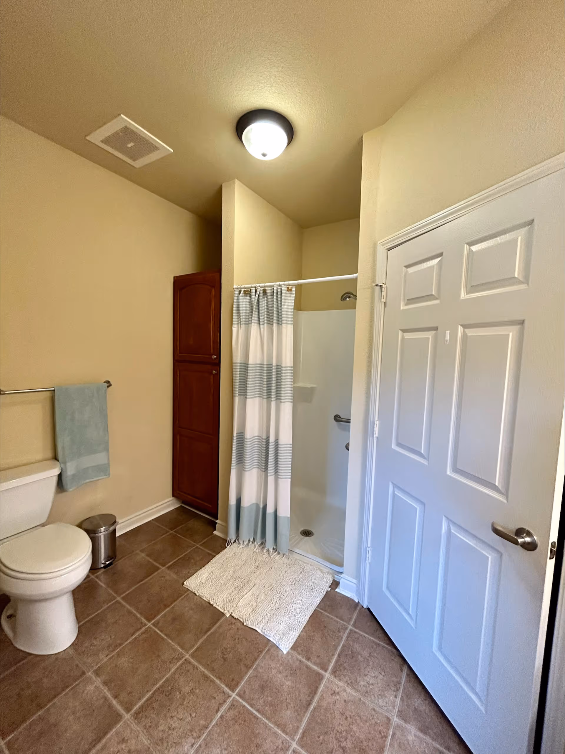 A bathroom with beige walls and brown tiled floor featuring a white toilet with a light blue towel hanging on a metal rack above it, a small silver trash can next to the toilet, a wooden cabinet, a shower with a white and light blue striped curtain, and a white door with a silver handle.