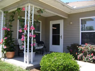 A covered porch area of a residential building with a white door and windows. The porch has white decorative metal supports with pink flowering vines climbing on them. There are two cushioned chairs on the porch, and the surrounding garden includes green bushes and flowering plants.