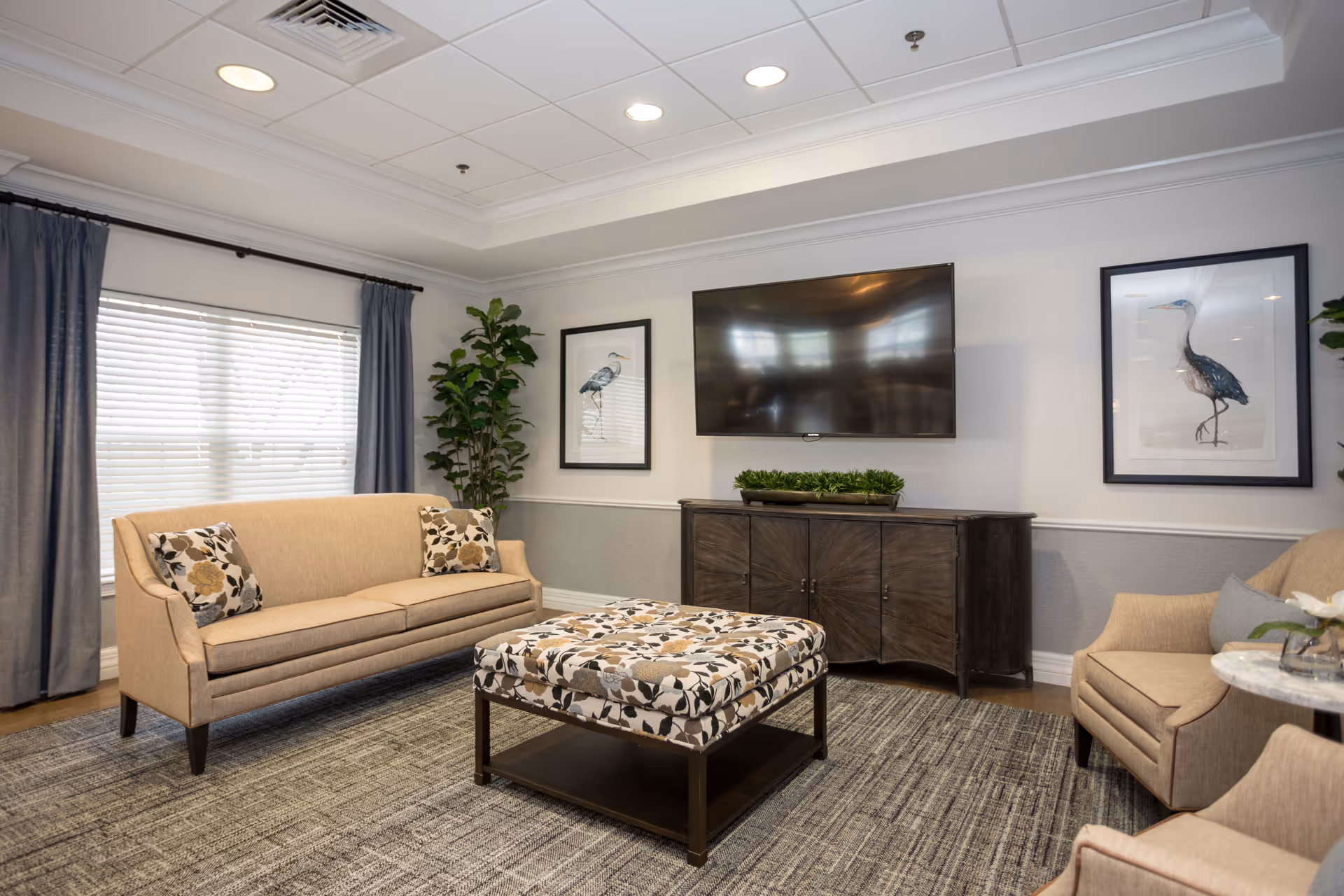 A bright common living room with a beige sofa, armchairs, patterned ottoman, wall-mounted TV and framed bird prints.