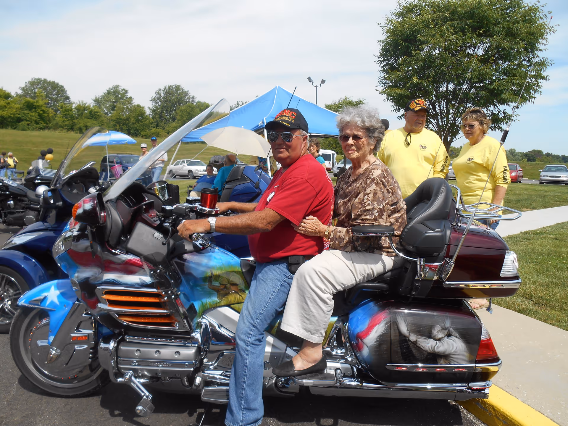 An elderly woman and a man sitting on a decorated motorcycle outdoors during a sunny day. The man is wearing a red shirt and a black cap with sunglasses, while the woman is dressed in a patterned blouse and light-colored pants. In the background, there are other people standing near a blue canopy tent and parked cars on a grassy area.