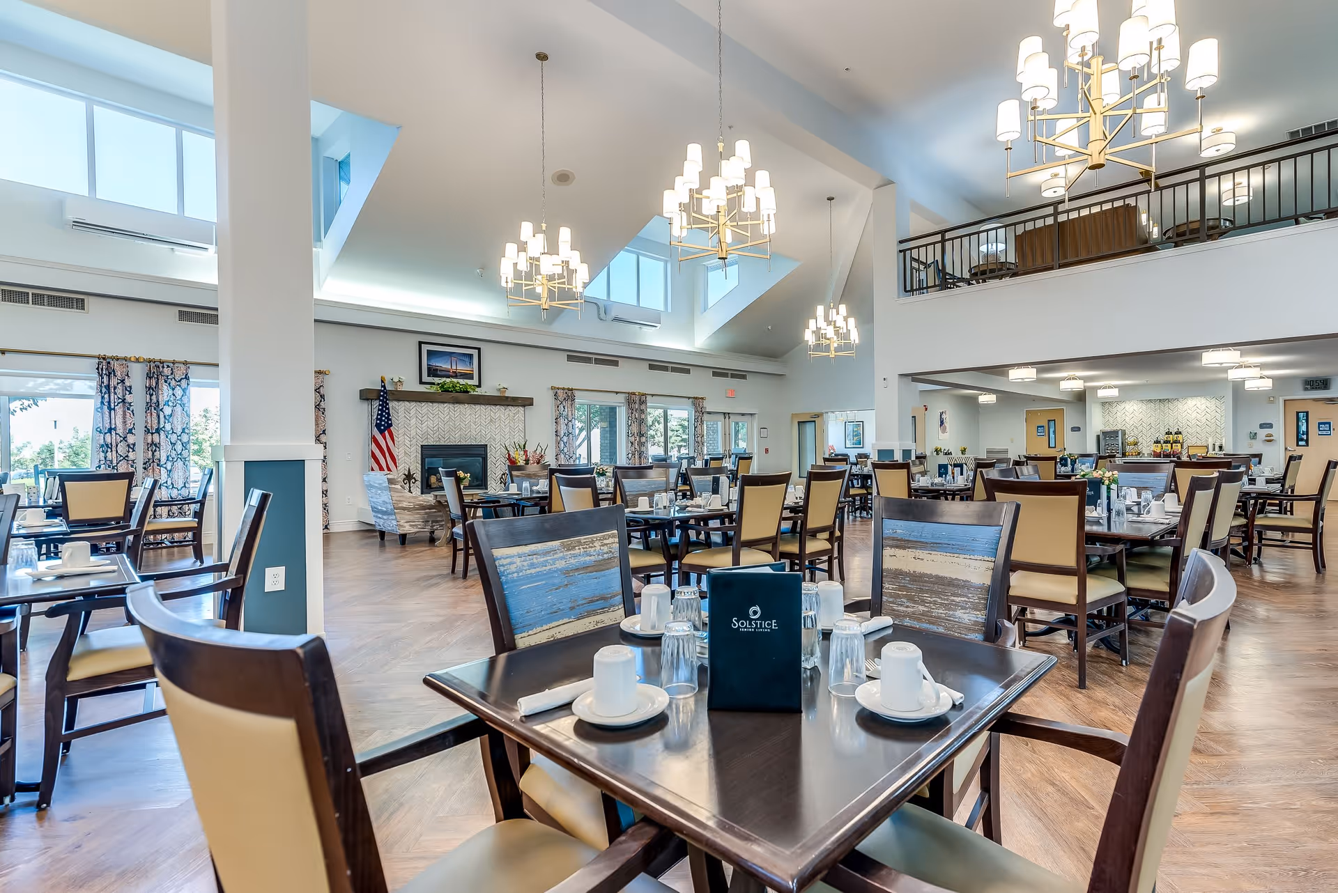 A spacious and well-lit dining room in a senior living facility with multiple wooden tables and chairs arranged neatly. The room features large windows with patterned curtains, a fireplace with an American flag beside it, and modern chandeliers hanging from a high ceiling with skylights. A menu with the name Solstice Senior Living is placed on one of the tables.