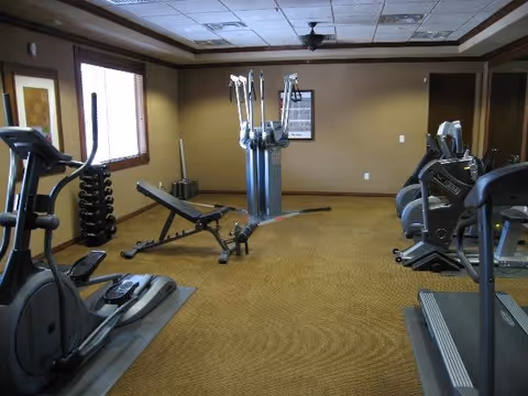 A fitness room with exercise equipment including a treadmill, elliptical machines, a weight bench, dumbbells on a rack, and a multi-station gym machine. The room has beige walls, a carpeted floor, and windows with blinds.