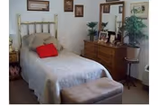 A bedroom with a brass bed frame, beige bedding, and a red pillow. There is a wooden dresser with a mirror, several framed pictures on the wall, a potted plant on a stand, and a cushioned bench at the foot of the bed.