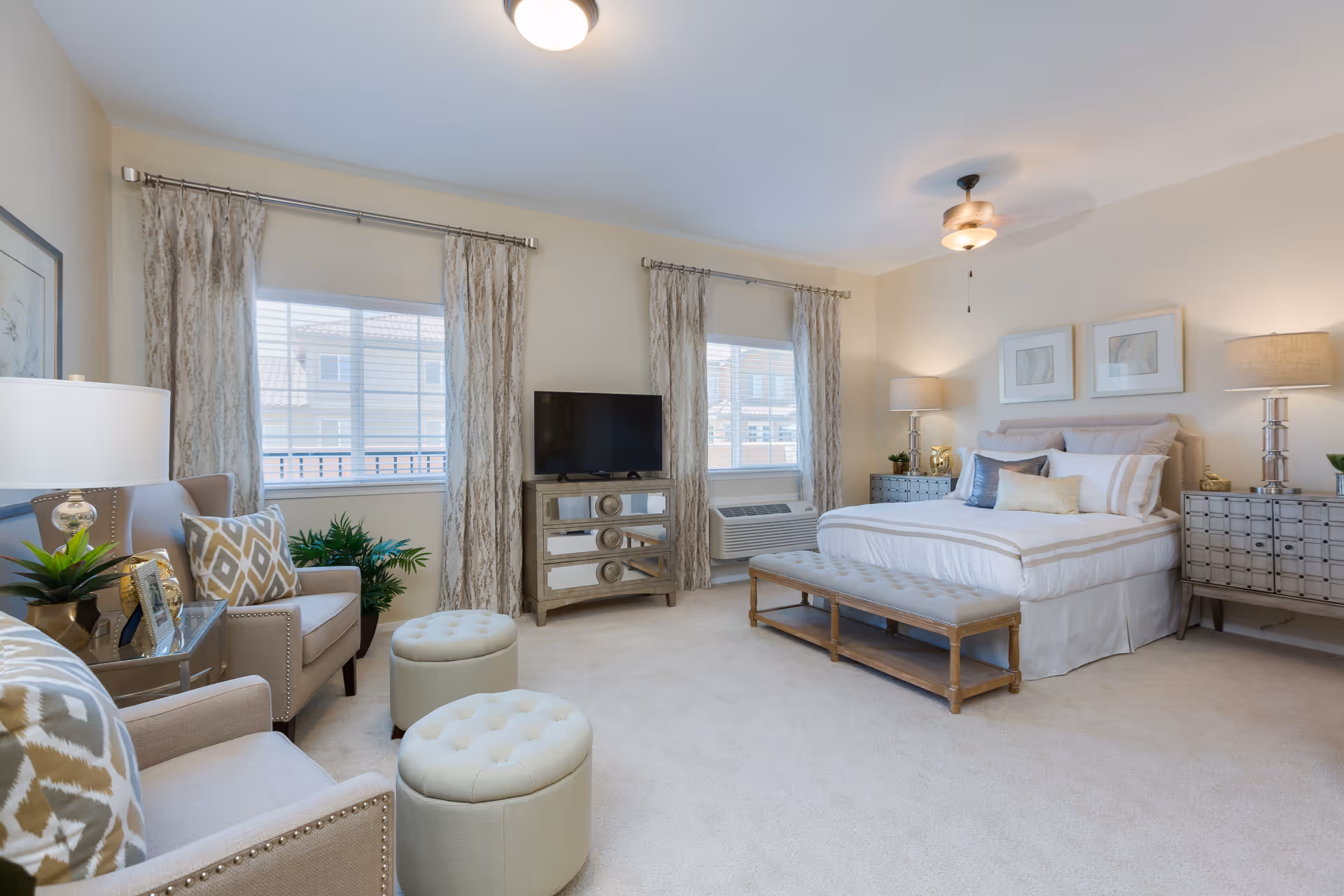 A bright and spacious bedroom in a senior living facility featuring a large bed with multiple pillows, two bedside tables with lamps, a bench at the foot of the bed, two armchairs with patterned cushions, two round ottomans, a TV on a mirrored dresser, and two windows with patterned curtains allowing natural light to fill the room.
