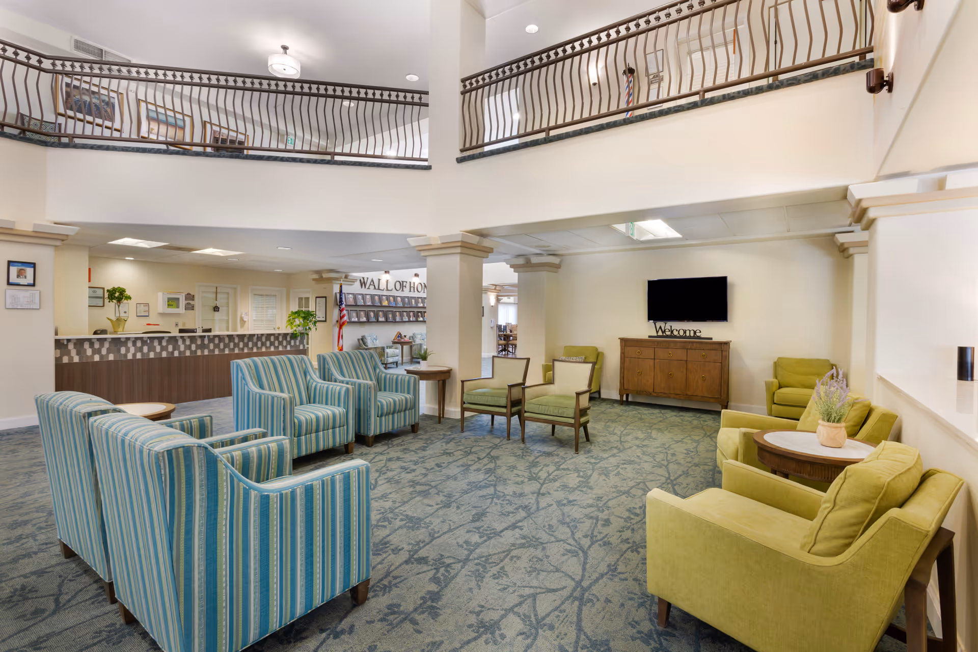 Spacious two-story senior living lobby with multiple striped and green armchairs arranged around tables, a reception desk, and a wall-mounted TV.