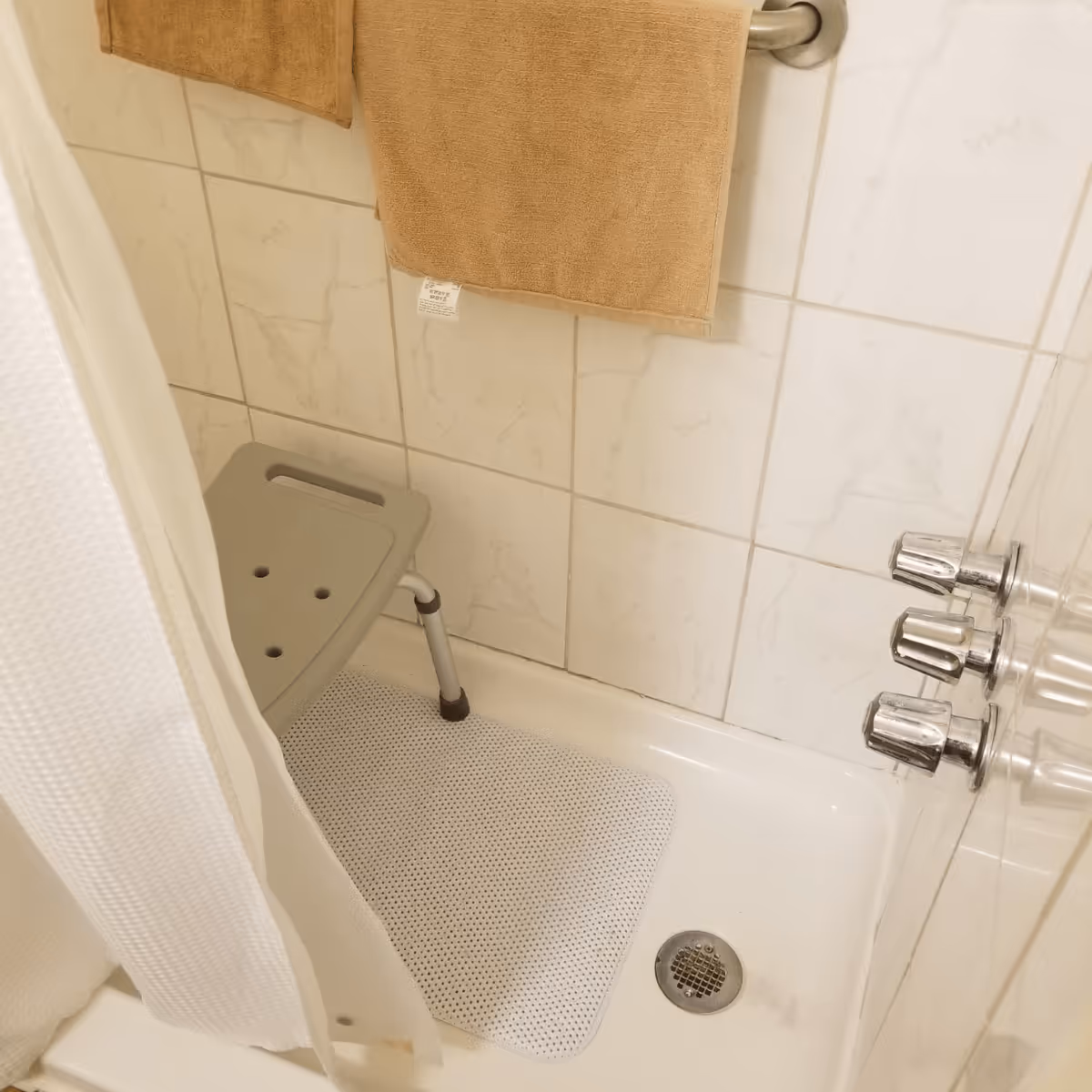 Bathtub shower area with a gray shower seat, white non-slip mat, beige towel hanging and chrome faucet knobs.