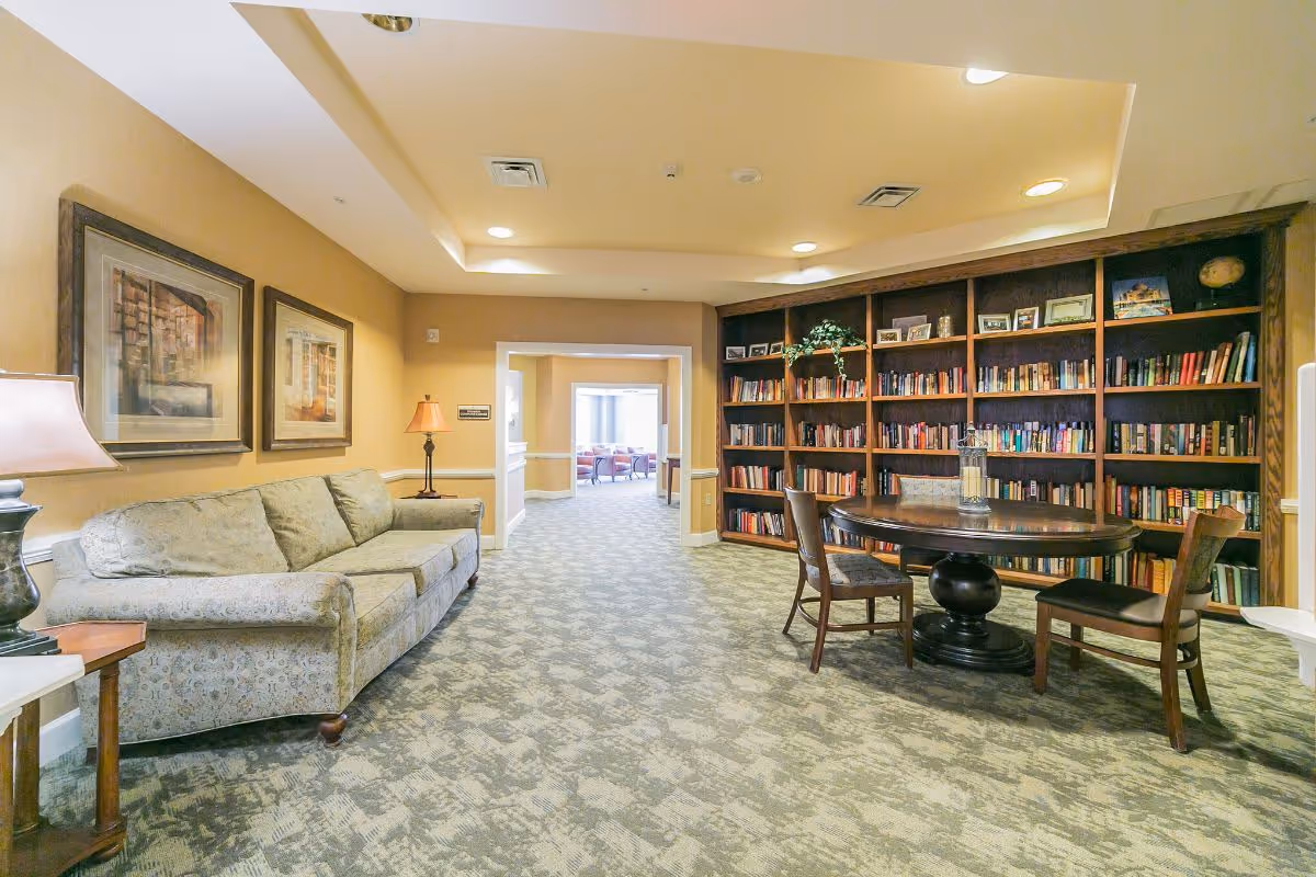 A cozy interior room with a patterned carpet, a beige sofa along the left wall, two framed pictures above the sofa, and a wooden table with three chairs in front of a large bookshelf filled with books and decorative items. The room is softly lit with ceiling lights and a table lamp, and there is an open doorway leading to another room with additional seating.